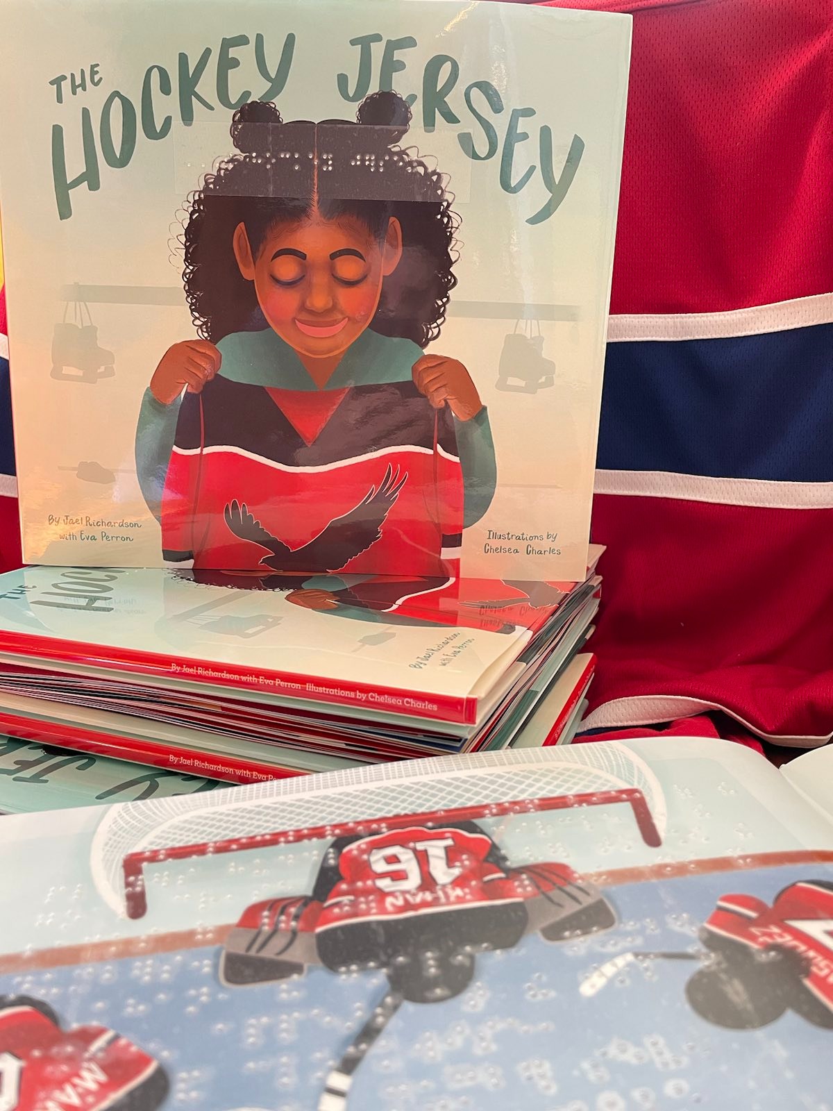 A children's book sits upright on a stack of the same books in front of a red and blue hockey jersey. The book cover has Kareema, a Black girl, standing in front of a washed-out locker room background, looking down at the hockey jersey she holds up to her chest. The hockey jersey is red and black with a black osprey on the chest. Green text “The Hockey Jersey” arches over Kareema’s head. The title, in braille, is below the title.